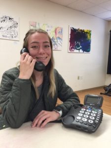 Mikaela holds the handset to her ear and the phone with large size numbers is on the table in front of her