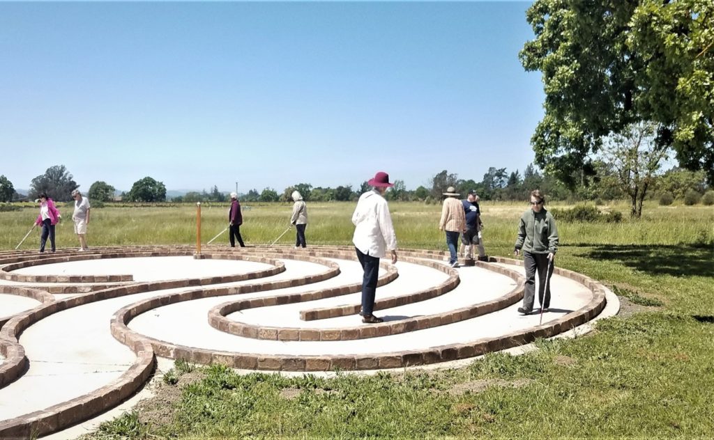 EBC Labyrinth outdoor walk. In the foreground, four of the participants are using their long white canes to guide themselves as they walk among others, following the concrete path. The Labyrinth’s pattern of swirling curves in the drying grass leads to the center where the wooden Peace pole orients them to easily find the way to the exit. On the horizon a variety of green trees make a dividing line against the blue sky.