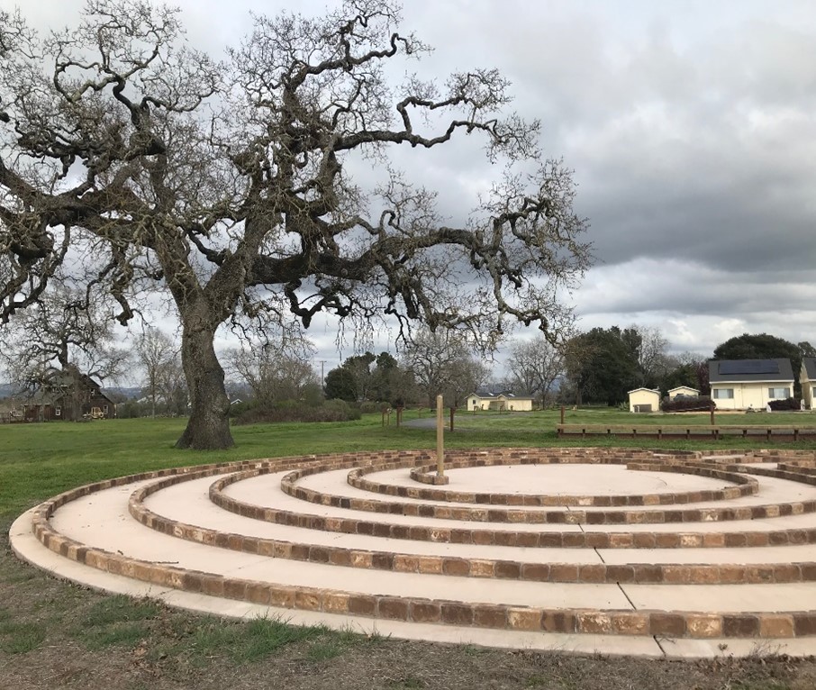 a winter day at the labyrinth has a sky full of rolling grey clouds. the giant oak tree without leaves, is behind the labyrinth on the left. in the far background are the yellow buildings of EBC.
